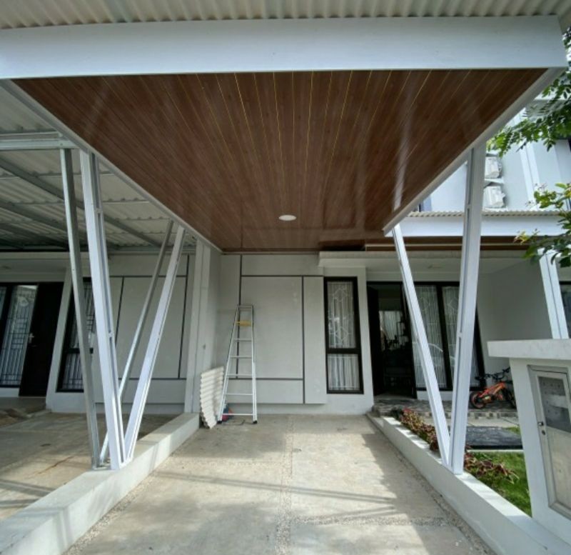 ampilan kanopi modern menggunakan atap Alderon putih dengan plafon PVC motif kayu, dipadukan dengan rangka besi hollow putih berbentuk V, terpasang di halaman depan rumah minimalis dua lantai.