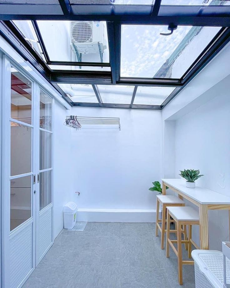 Area indoor minimalis dengan atap kanopi sliding manual berbahan kaca dan rangka besi hitam. Ruangan berwarna putih dengan meja dan kursi kayu, serta tanaman hias sebagai dekorasi.