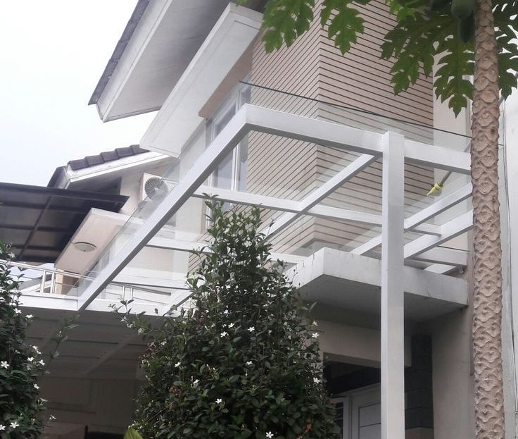 Kanopi kaca tempered pada rumah dua lantai dengan rangka putih dan taman hijau tropis di bagian depan.