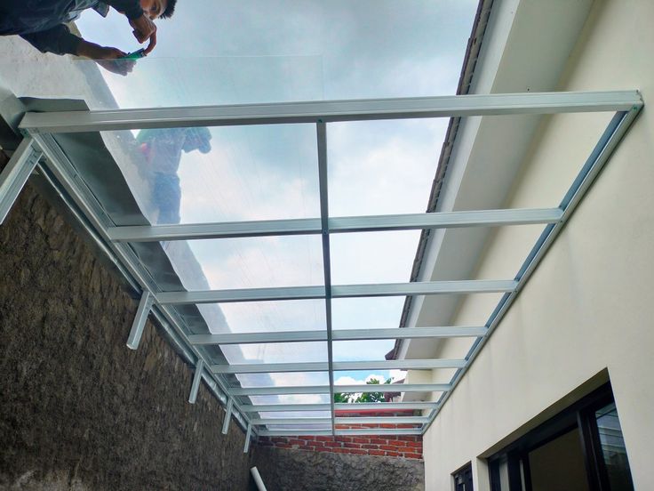 Pemasangan kanopi kaca tempered transparan dengan rangka besi hollow putih di area samping rumah, memberikan pencahayaan alami dan tampilan modern.