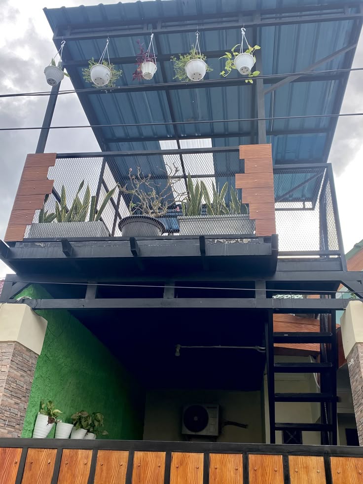 Kanopi balkon dengan atap baja ringan berwarna biru dan struktur besi hitam, dilengkapi pagar kawat, pot tanaman hias gantung, serta tangga besi samping.