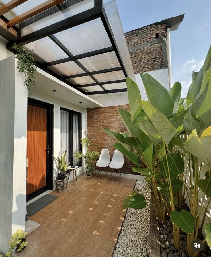 Teras rumah minimalis modern dengan kanopi polycarbonate bening, rangka besi hitam, dilengkapi kursi putih dan tanaman tropis yang rindang di sisi halaman.