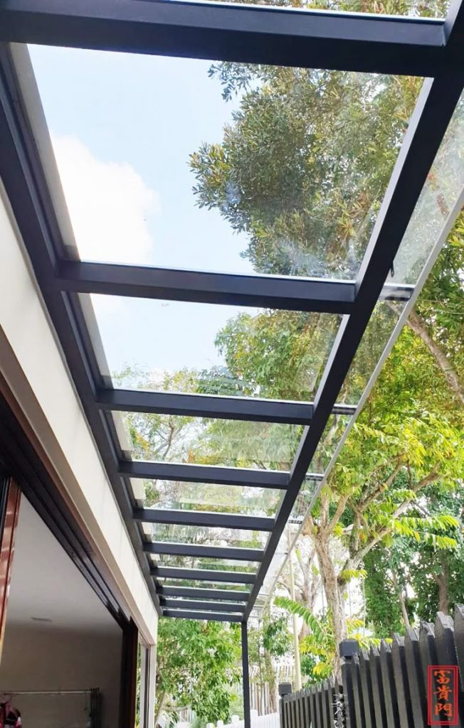 Kanopi kaca tempered transparan dengan rangka besi hitam, terpasang di sisi rumah modern dengan latar pepohonan rindang