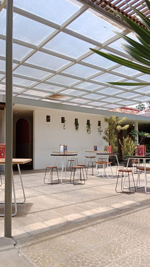 Kanopi polycarbonate transparan dengan rangka besi putih berbentuk grid, terpasang di area outdoor kafe modern yang terang dan sejuk.
